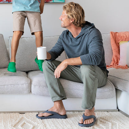 Male model wearing Ara Thong Sandal in mineral watching kids jump on the sofa while sipping coffee
