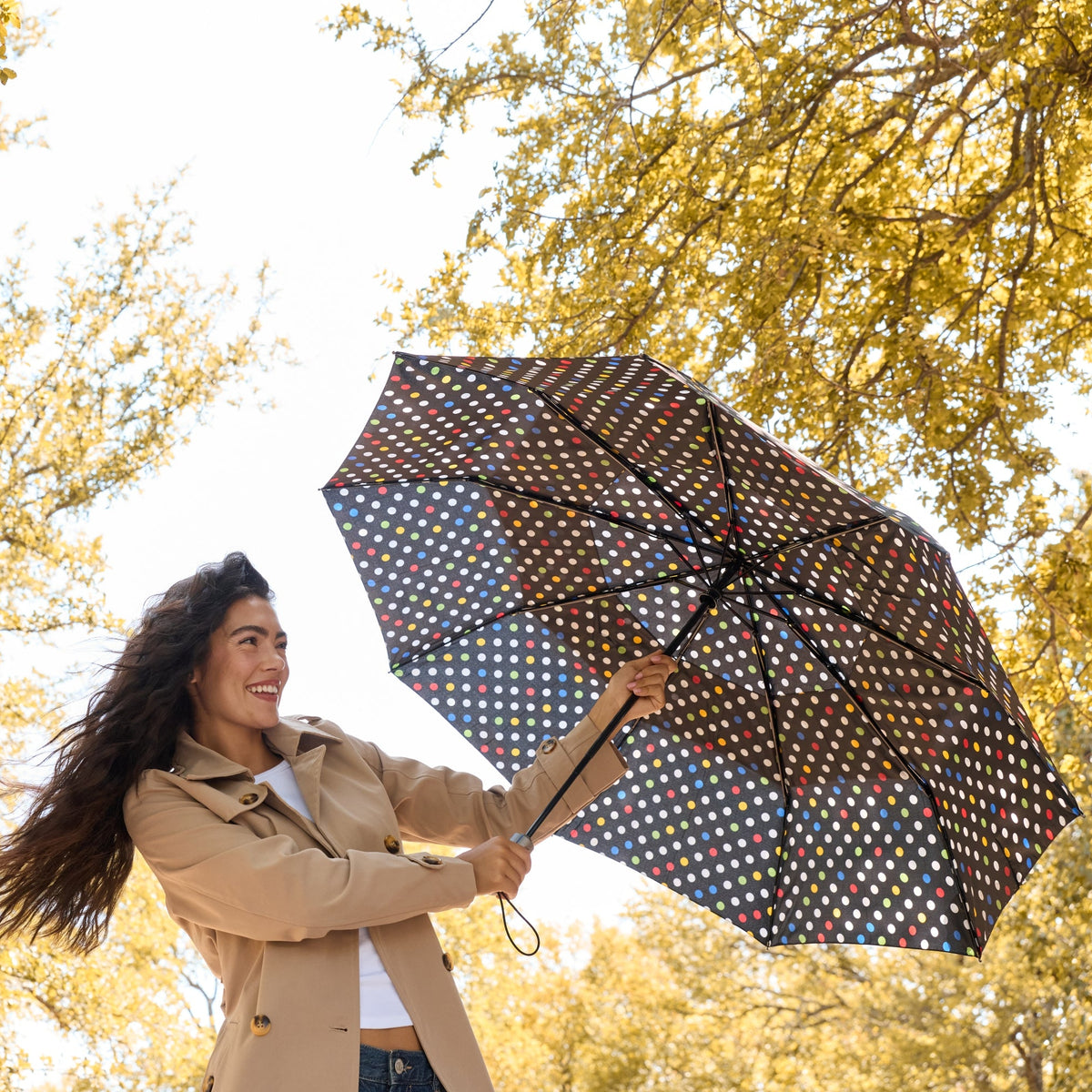 A woman in a beige trench coat holds up a black umbrella with a colorful polka-dot pattern in an outdoor park with golden autumn trees.

#color_blue